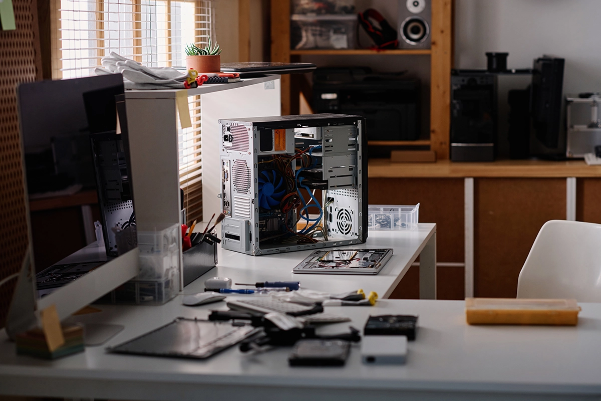 Open desktop computer case on a workbench surrounded by tools and computer parts in a repair workshop