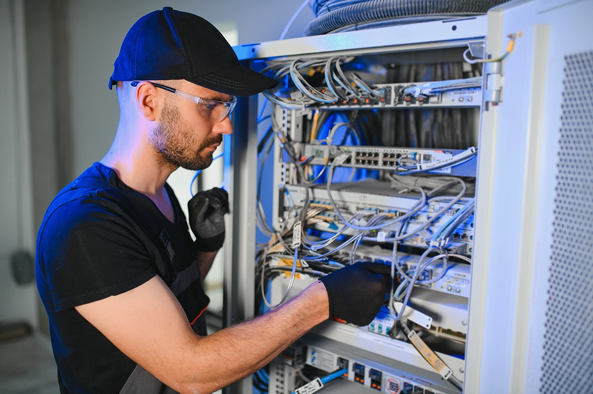 Man Working on Server - Computer Headquarters IT technician wearing gloves and cap maintaining network cables inside a server rack in a data center.