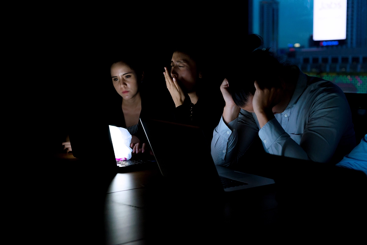 Office workers sitting in the dark during a power outage, illuminated by laptop screens and emergency light.