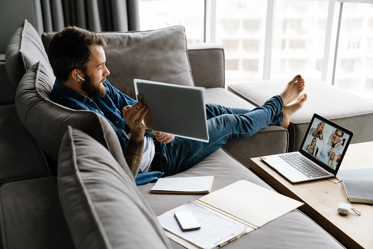 Man working remotely from home, using a laptop and tablet for a video conference call with colleagues.