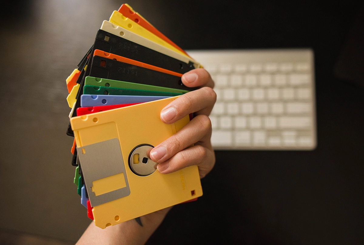 Person holding a fan of colorful floppy disks in front of a computer keyboard, symbolizing outdated data storage technology compared to modern cloud solutions.