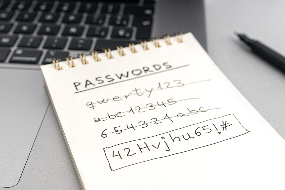 Notebook labeled “passwords” showing a complex alphanumeric password, placed beside a laptop to represent strong password management.