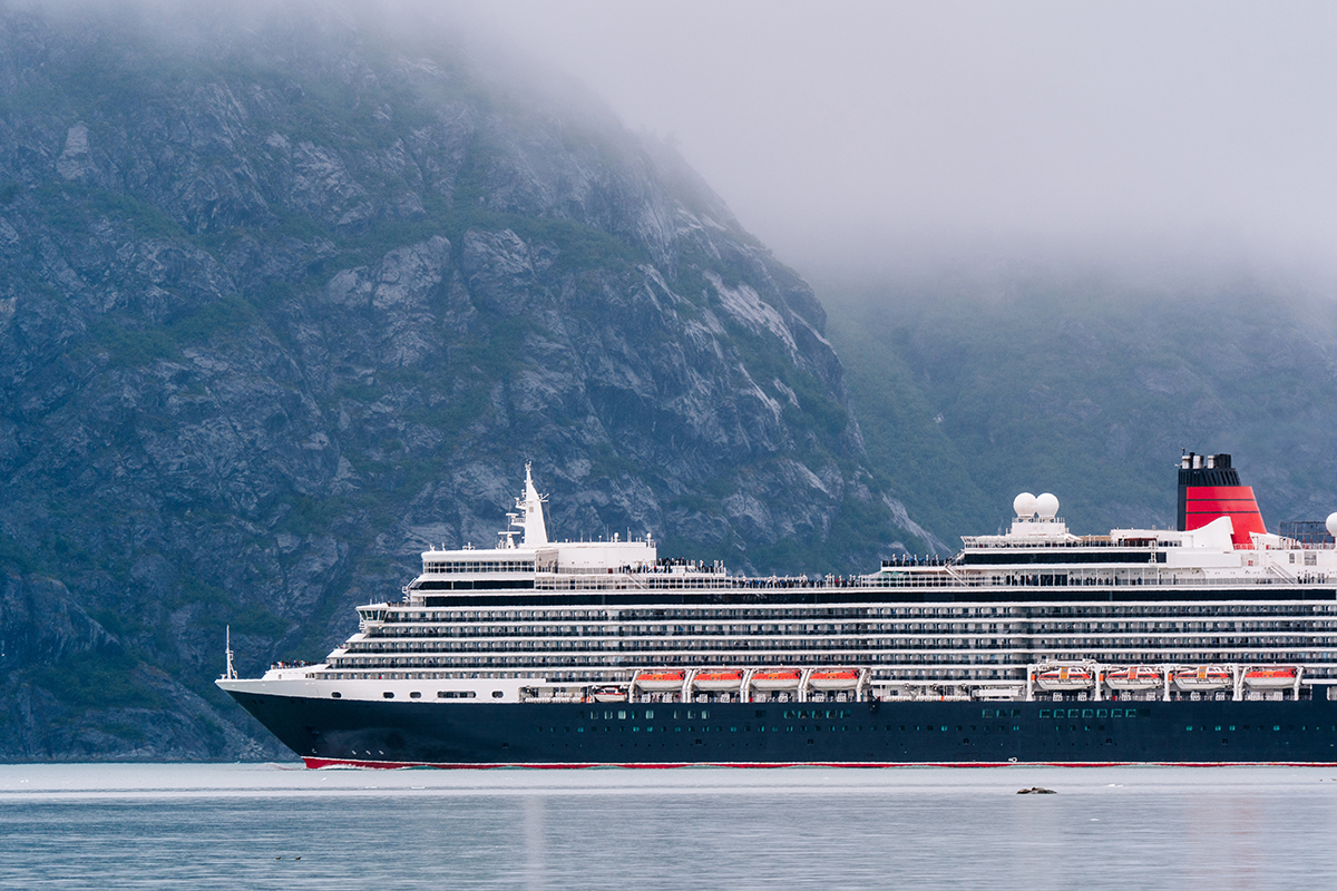 Large cruise ship sailing through misty Alaskan fjord with steep green mountains and calm water