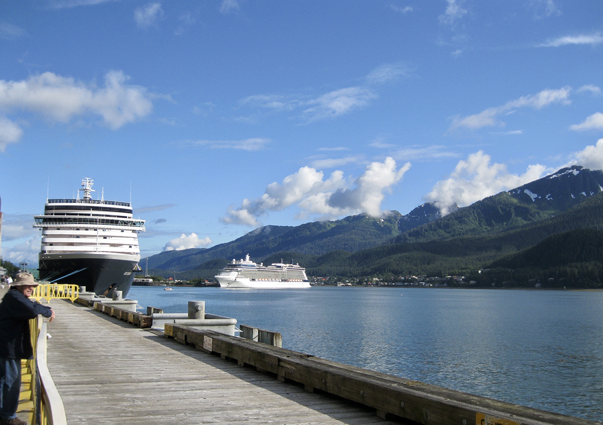 Two large cruise ships docked at a scenic mountain port with blue sky, waterfront pier, and coastal travel tourism destination.