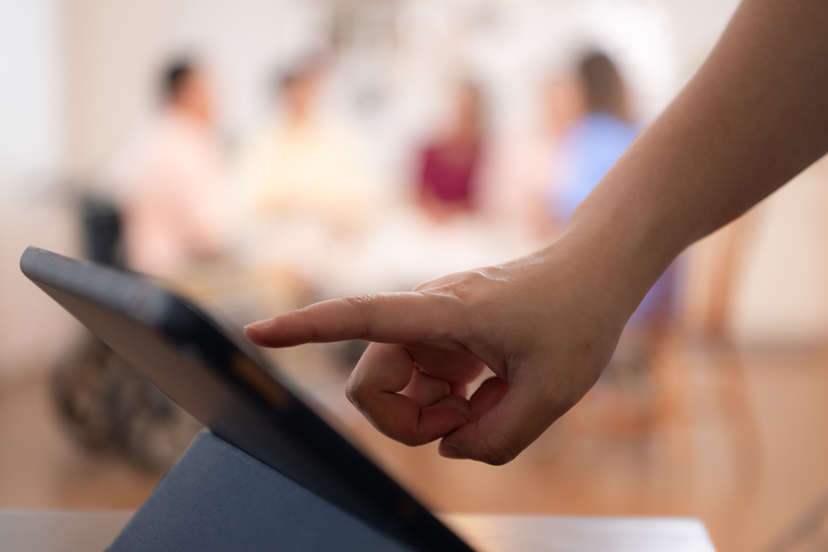 Hand tapping a tablet in an office or meeting room, with blurred people in the background.