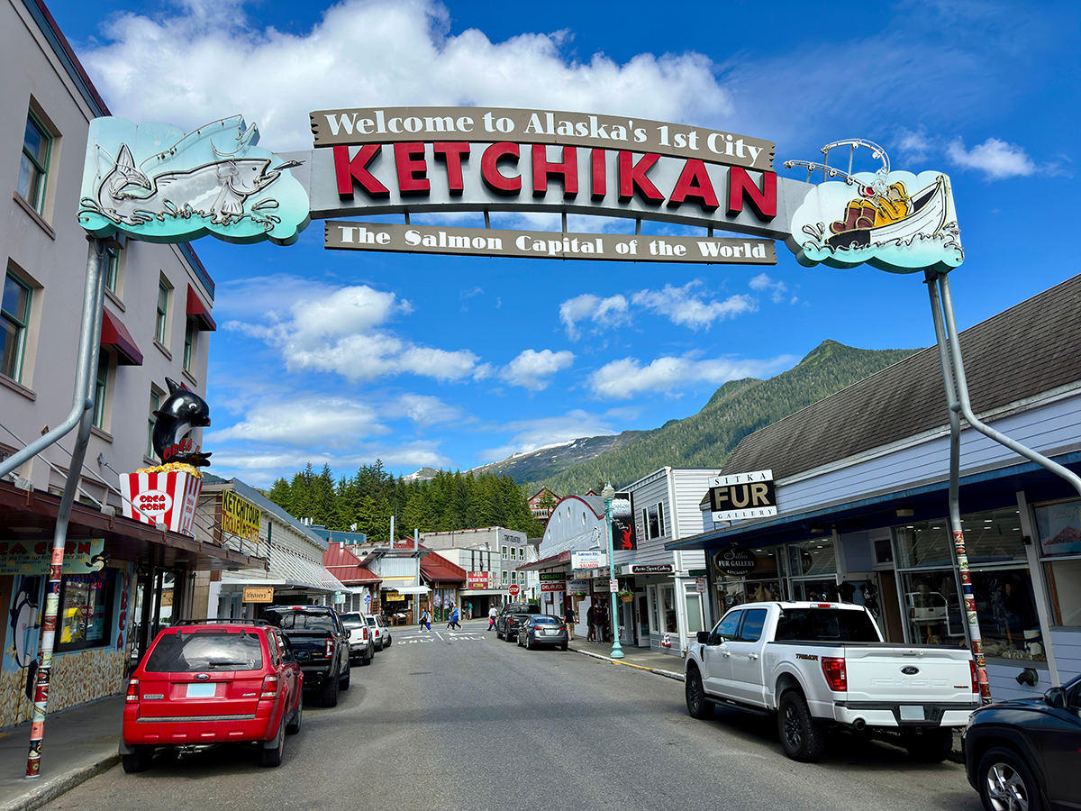 Street view of the Ketchikan welcome sign in Alaska, with shops, cars, mountains, and a blue sky in the background.