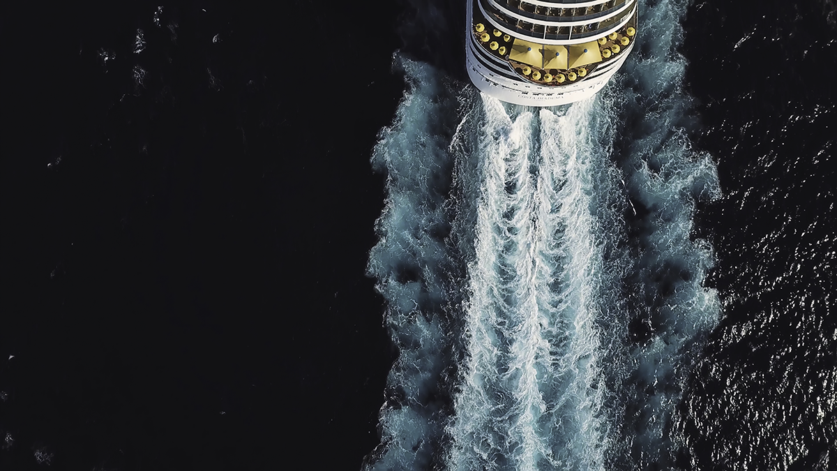 Aerial view of a cruise ship sailing through dark ocean water, leaving a wide turquoise-and-white wake behind it.